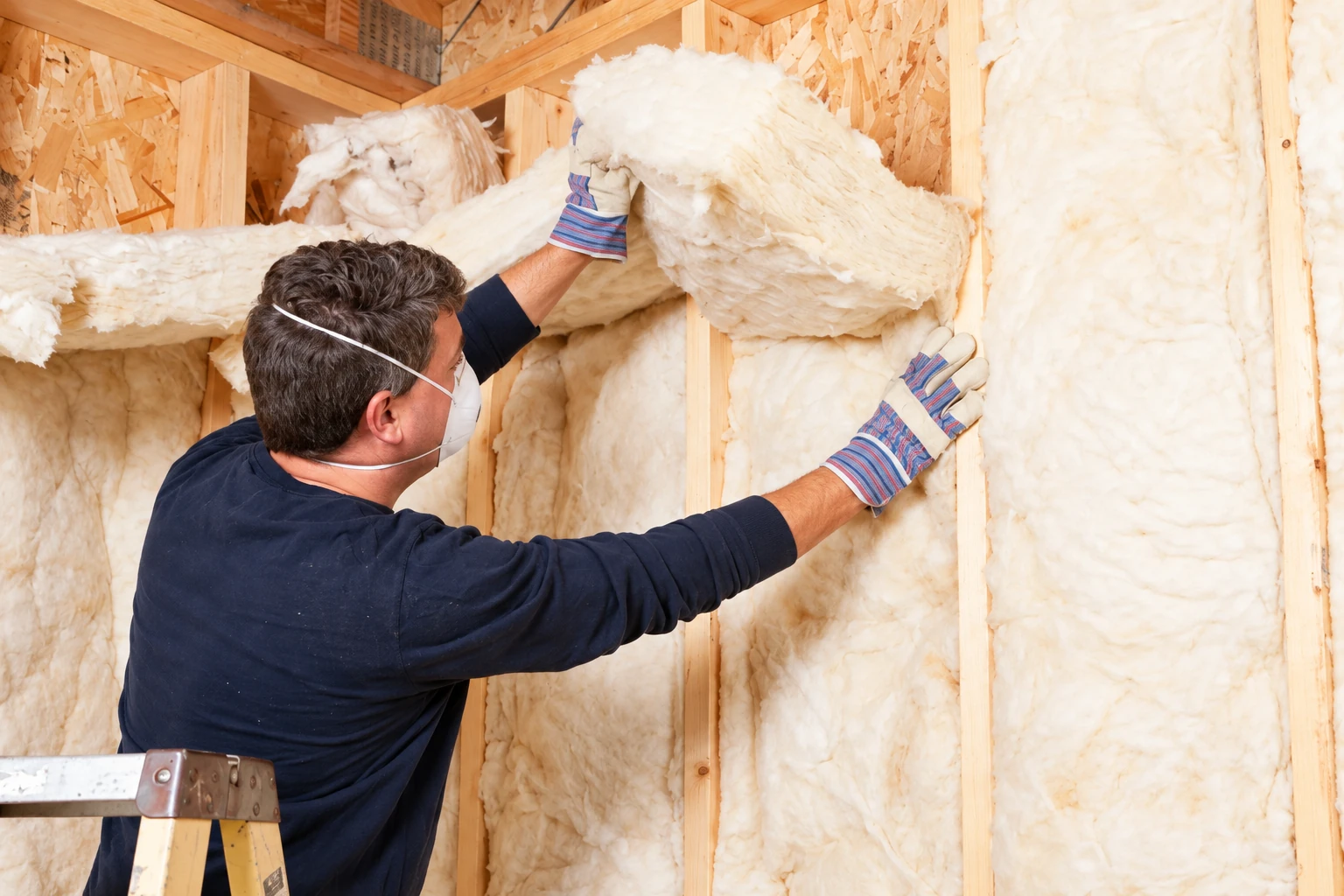 A worker in a mask and gloves fits fluffy fiberglass insulation between the exposed studs of a house wall.