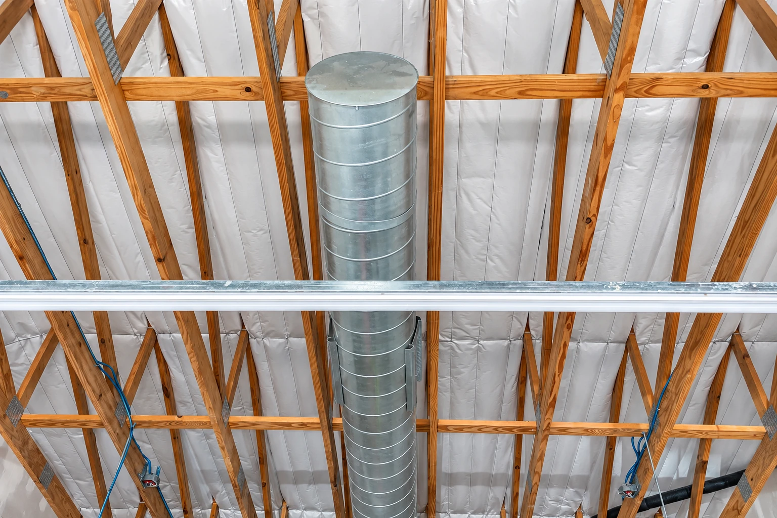 White vapor barrier installed between exposed wooden roof trusses around a large metal ventilation duct.