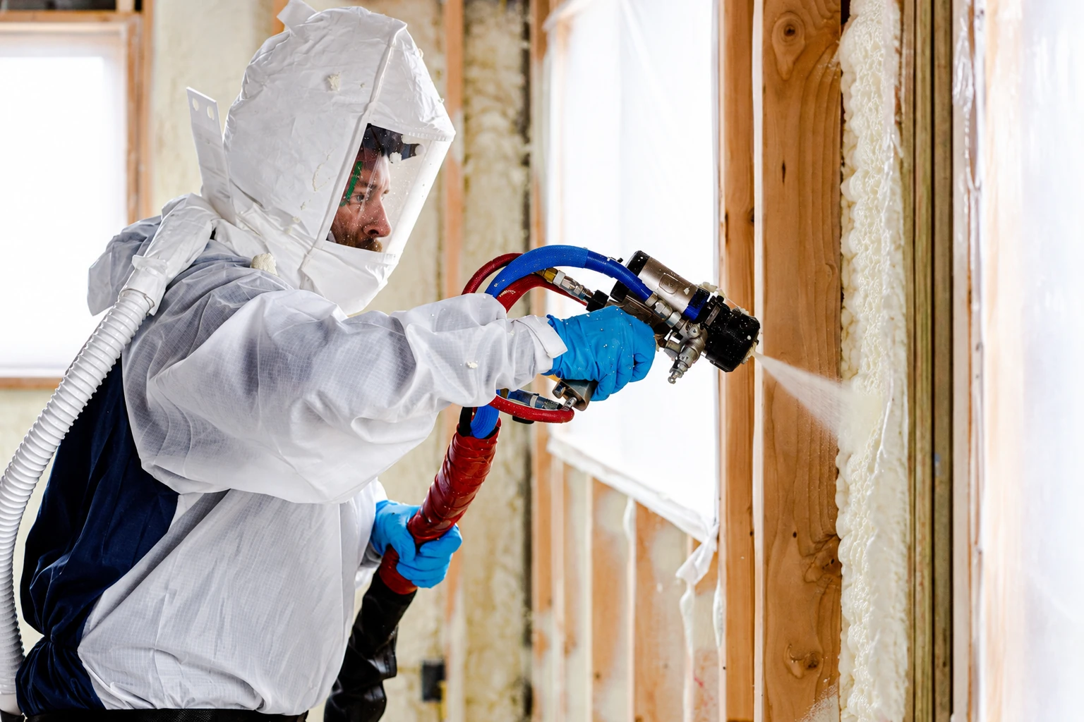 Insulation worker in protective gear sprays foam into the spaces between wooden wall studs.